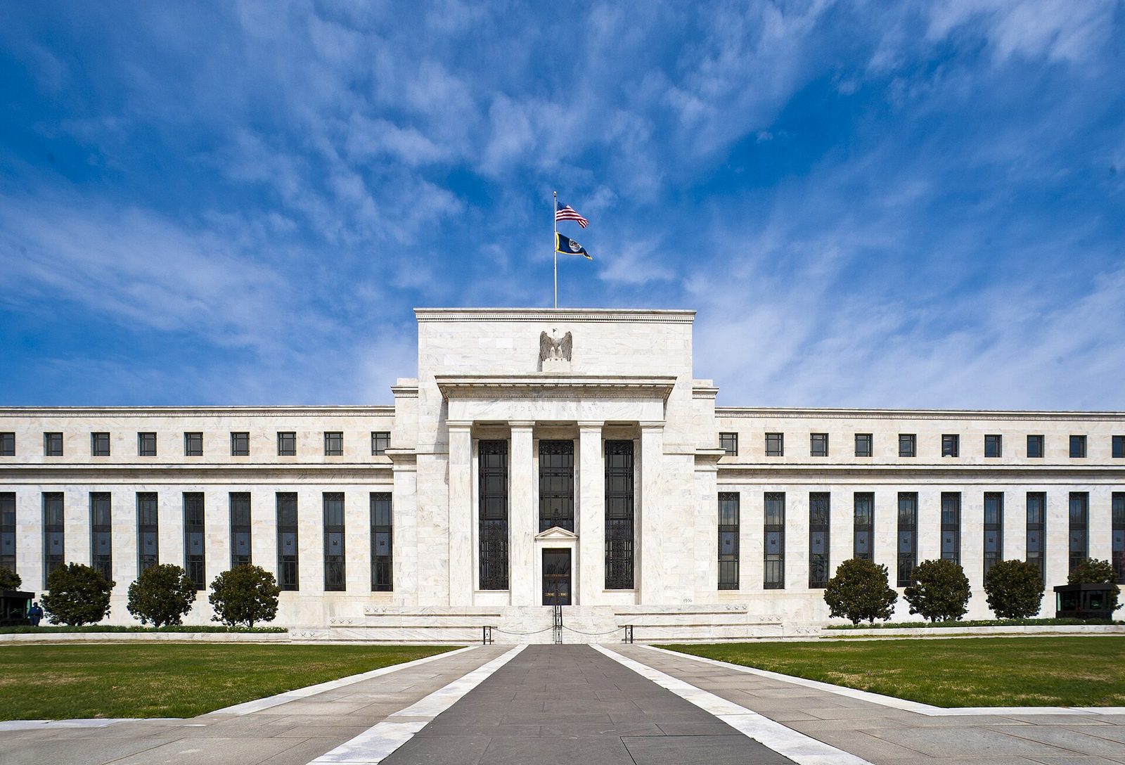 The white marble front of the Marriner S. Eccles Federal Reserve Board building in Washington under a blue sky, with a stone eagle over the central entrance and an American flag on the roof