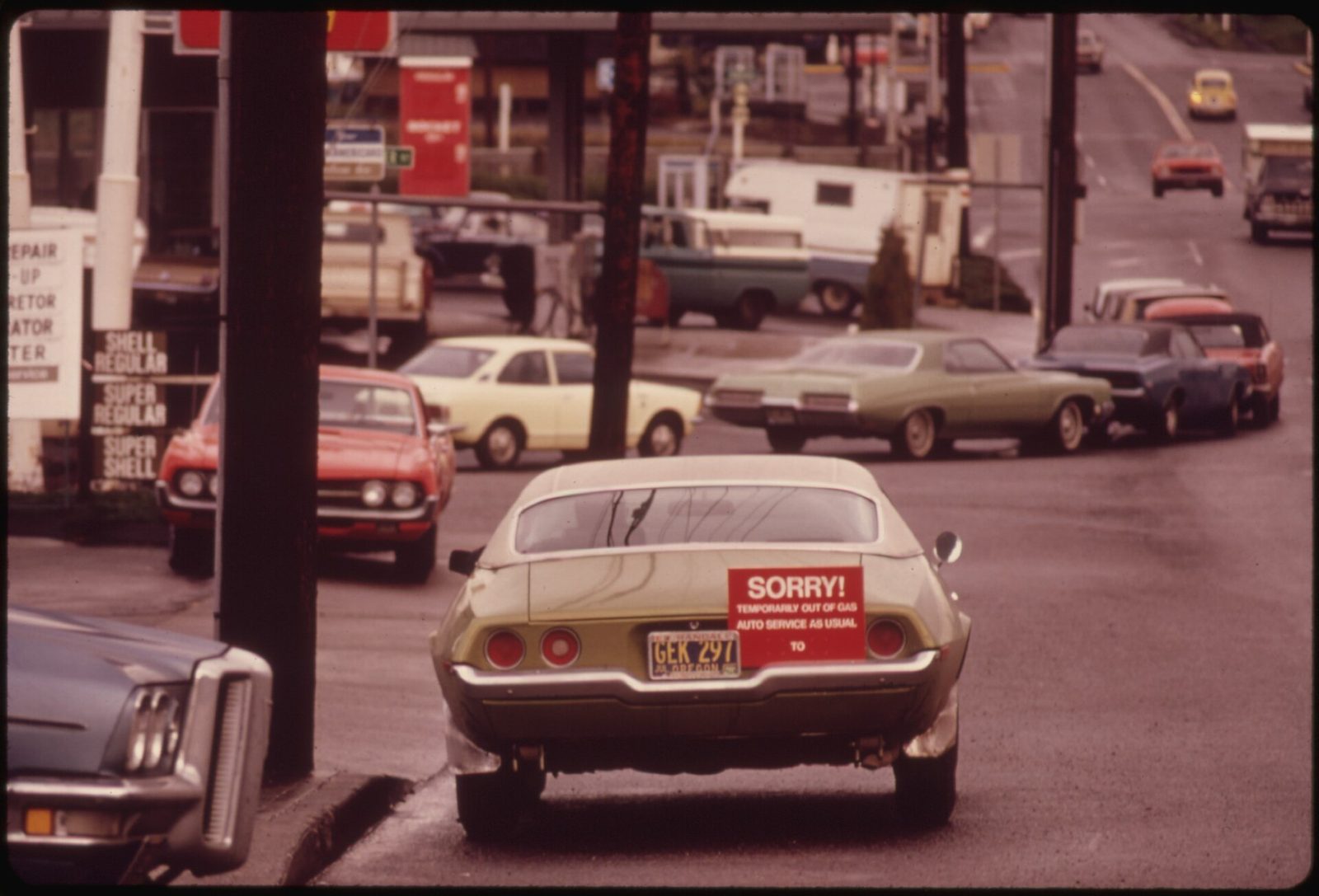 A 1973 colour photograph looking down a wet Portland street at a gas station: a green-gold Chevrolet with an Oregon plate has a red sign in its rear window reading SORRY! TEMPORARILY OUT OF GAS, AUTO SERVICE AS USUAL, with a Shell price board and a queue of cars behind